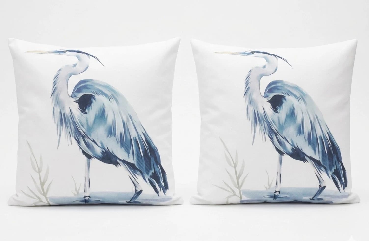 White coastal throw pillows featuring blue heron and black feather pattern, standing on white water with green reeds.